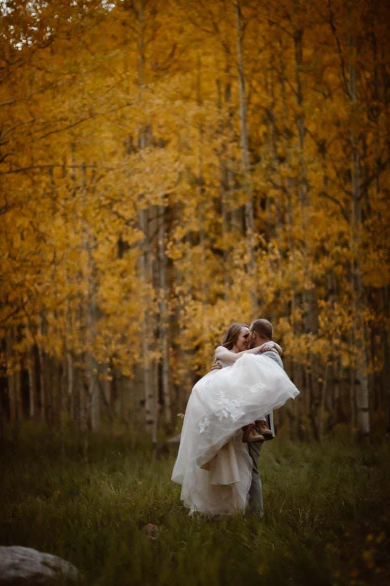 Autumn soaked Maroon Bells Wedding in Aspen where the newlyweds are surrounded by golden colors of Aspe trees. The groom is picking up the bride in her bridal gown and they are standing in the embrace of love.