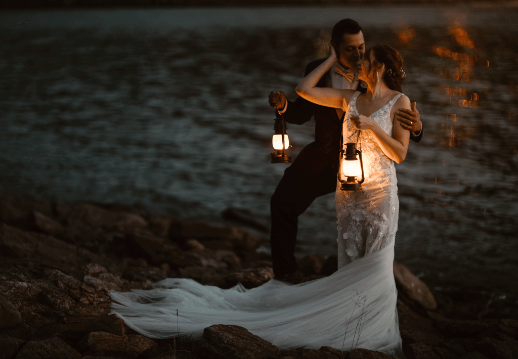 “Romantic Colorado elopement photographer captures an intimate lakeside wedding moment as the bride in a lace gown and the groom embrace while holding glowing lanterns. Set against a mountain lake at sunset, this photo highlights authentic love and adventure.” Twilight Vows in Estes Park Colorado Colorado Elopement Photographer