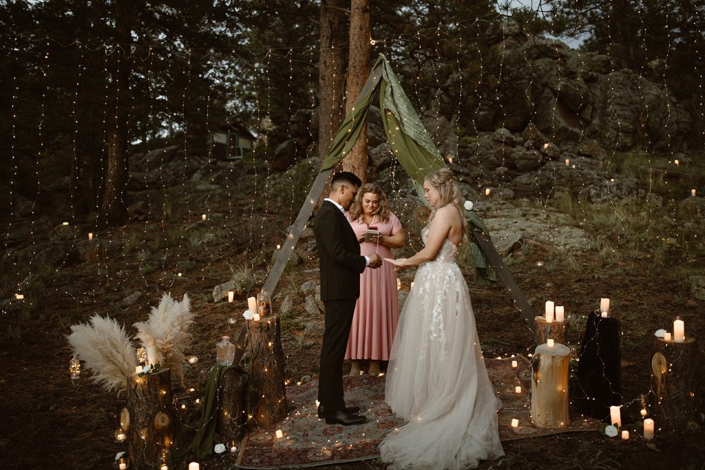  “Colorado elopement photographer captures an intimate forest wedding ceremony with bride and groom exchanging vows under a boho teepee decorated with candles, pampas grass, and fairy lights.” “Colorado elopement photographer captures an intimate forest wedding ceremony with bride and groom exchanging vows under a boho teepee decorated with candles, pampas grass, and fairy lights.”