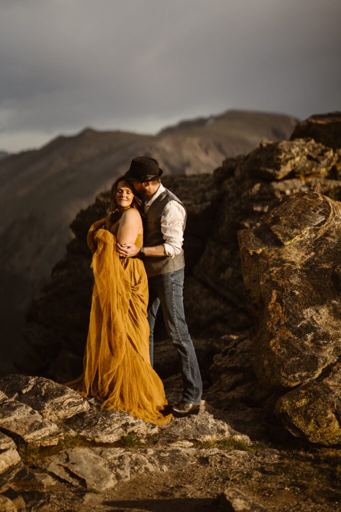 Couple cuddling on a rocky mountainside, woman in mustard yellow dress resting in partner’s arms, intimate Colorado elopement photography.