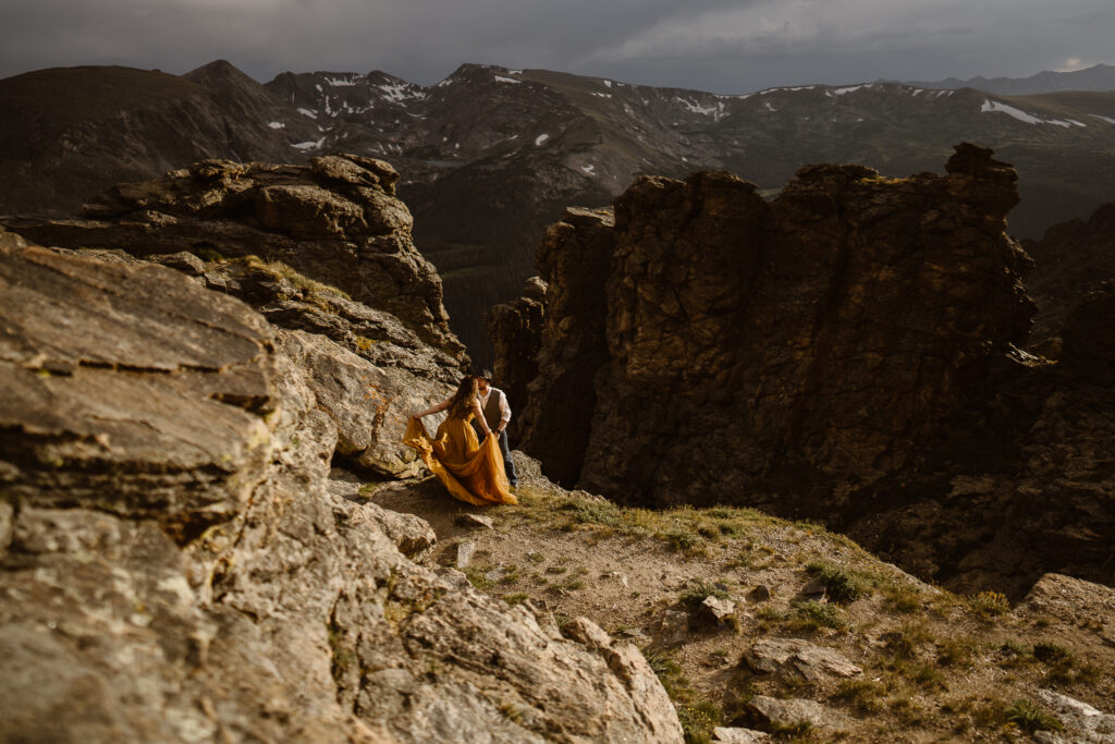 Romantic mountain elopement scene, couple holding hands and relaxing on rugged terrain, golden dress glowing in natural light, Colorado wedding photography.